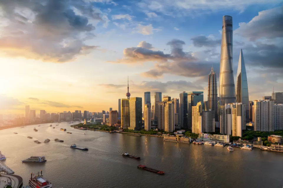 A view of Downtown Shanghai's skyscrapers, rising above a Huangpu River full of boats leaving wakes visible in the fading light of sunset.